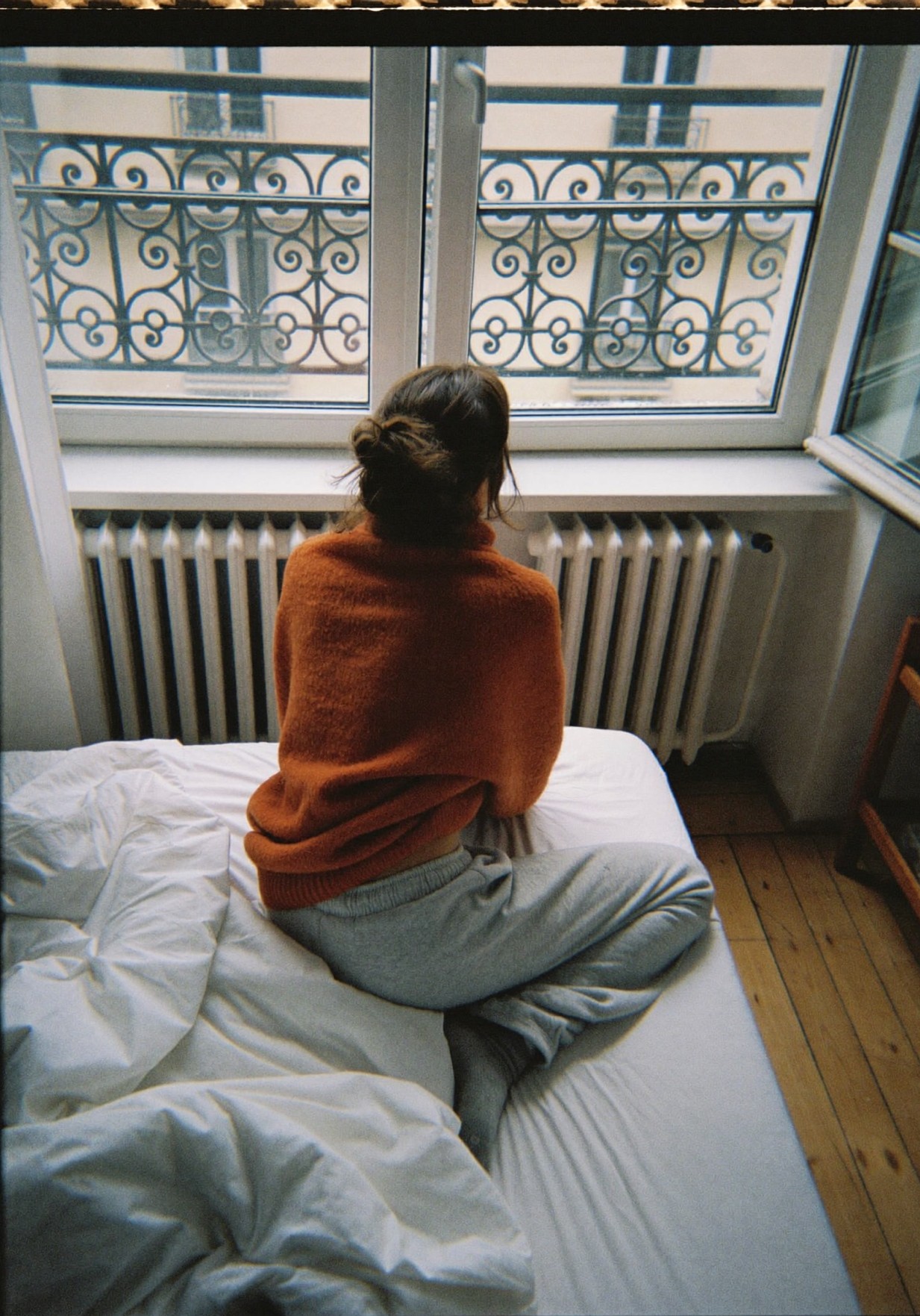 Femme de dos assise sur son lit, face à la fenêtre, enveloppée dans un gilet